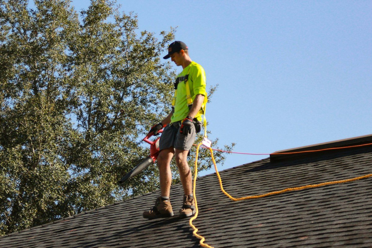 A roofer in a safety harness clearing debris from a shingle roof