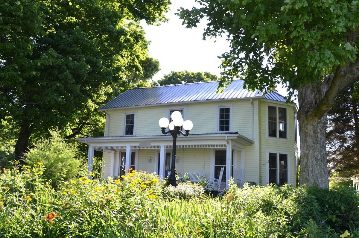 A pale yellow two-story house with a metal roof behind a summer garden